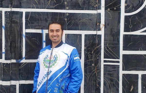 A man stands in front of a wall wearing a blue jacket.
