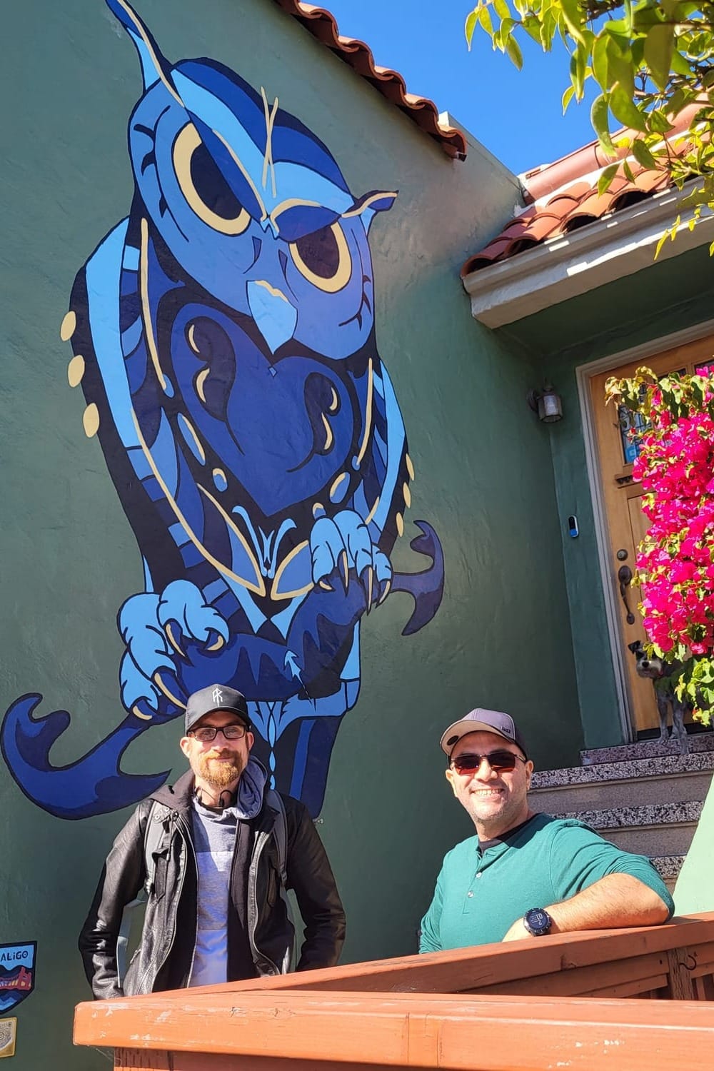 Two men in front of a mural of a blue owl on a house.