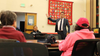 A man stands before a table with people seated in a meeting room.