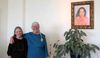 Two women stand beside a plant and a framed portrait.