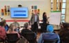 Two people stand and write down ideas on a board from a seated crowd.